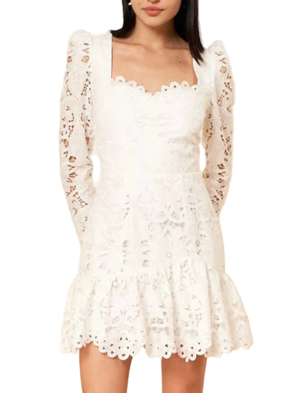 A woman wearing a white eyelet lace mini dress with long sheer sleeves, a sweetheart neckline, and a ruffled hem, shown against a light beige wall.