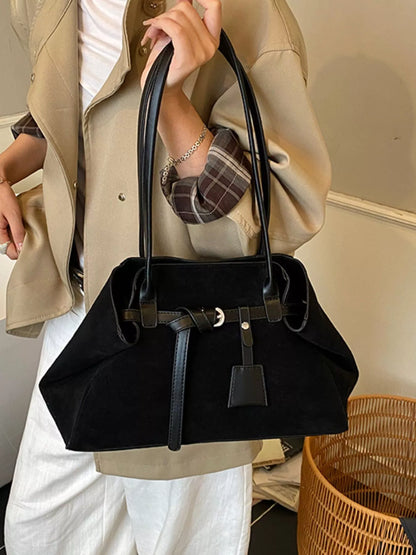A person wearing a beige blazer and white pants holds a black suede shoulder bag with black leather handles and a front strap detail in an indoor setting.