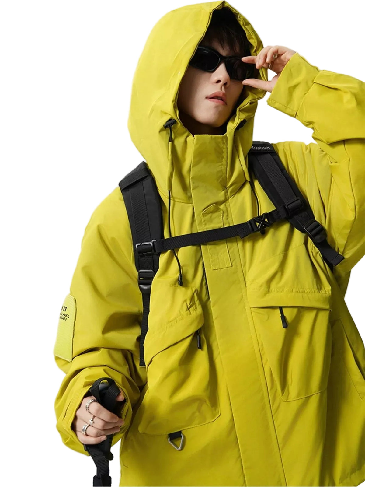 A person wearing a bright yellow hooded outdoor jacket with black backpack straps visible, black sunglasses, and holding a black handle against a light gray studio background.