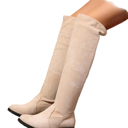A pair of knee-high beige suede boots worn on a model's legs, set against a rustic background of straw bales, a wooden stool, and a wooden floor.