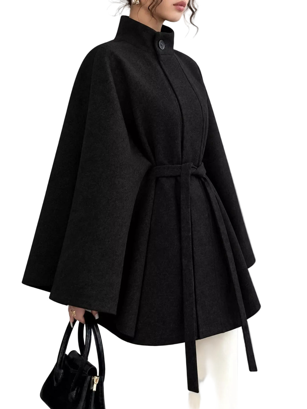 A woman wearing a black cape-style coat with a high collar, long wide sleeves, and a self-tie belt, paired with white pants and holding a black handbag against a plain gray studio background.