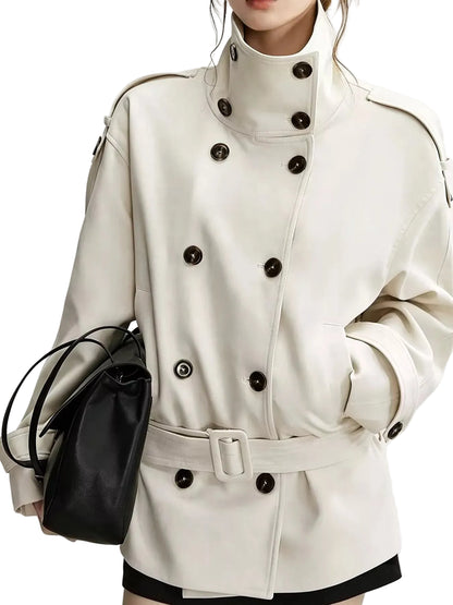 A close-up of a model wearing a cream-colored double-breasted trench coat with dark buttons and a matching belt, holding a black leather handbag against a plain light background.