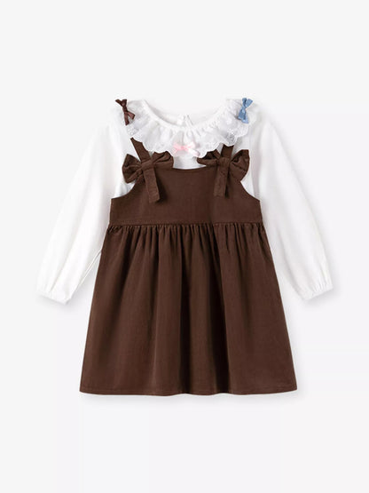 A toddler girl's long-sleeve dress featuring a brown pinafore-style skirt with shoulder bows over a white long-sleeve top, accented with a ruffled collar.