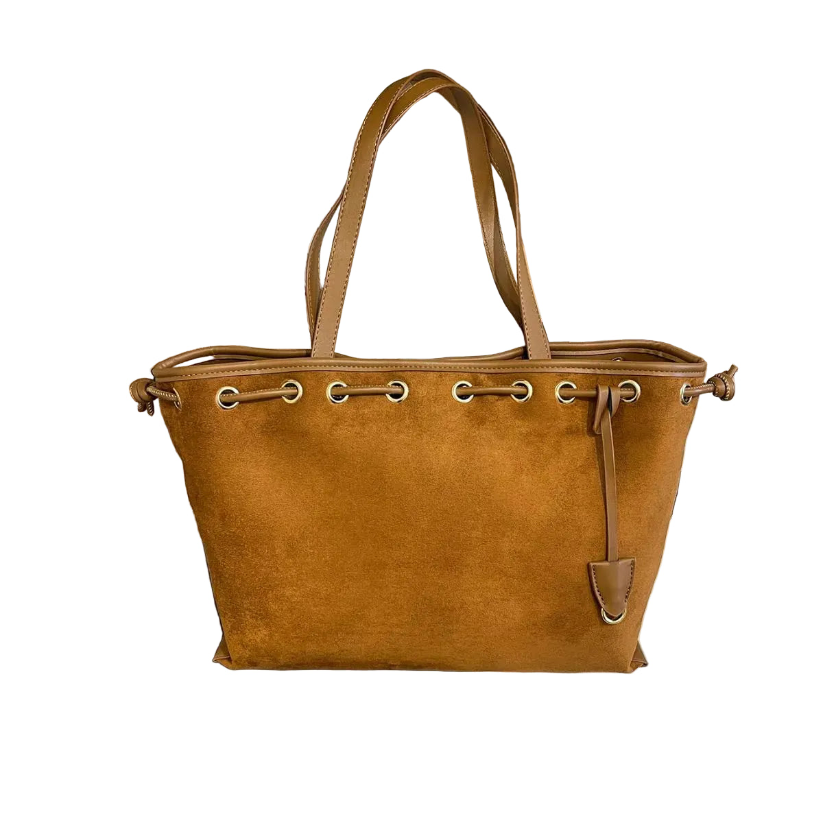 A tan suede tote bag with double shoulder straps and silver-tone grommets, resting on a black leather chair in front of a light wood bookshelf filled with books.