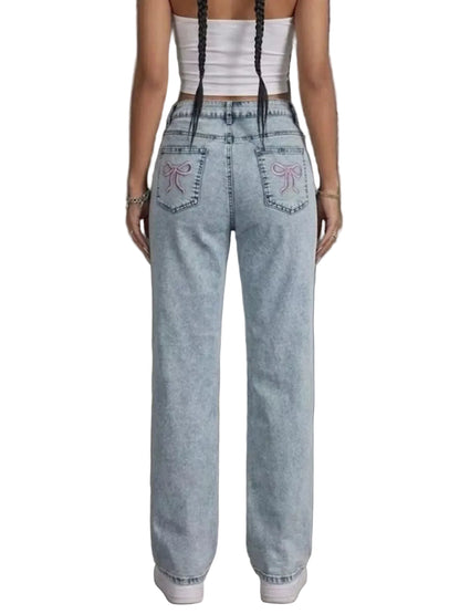 A person shown from the back wearing light-wash wide-leg jeans with pink bow embroidery on the back pockets, a white ribbed crop top, and white sneakers in an industrial indoor setting.