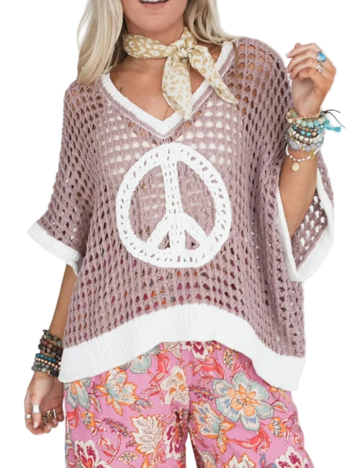 A woman wearing a loose, open-knit mauve-pink V-neck top with a large white peace sign motif on the front, paired with pink floral pants and a beige patterned neck scarf, against a plain light background.