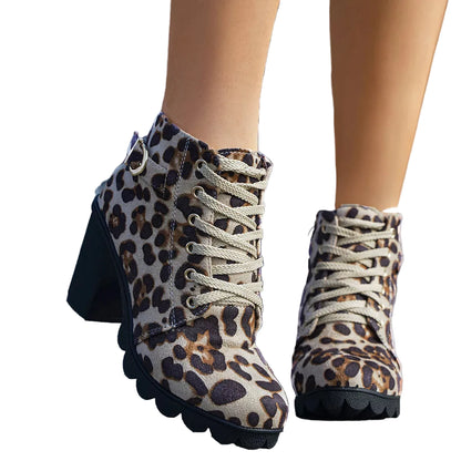 A pair of women's ankle boots with a beige, brown, and black leopard print pattern, featuring beige laces and chunky black heels, shown on a model standing on gray stone steps.