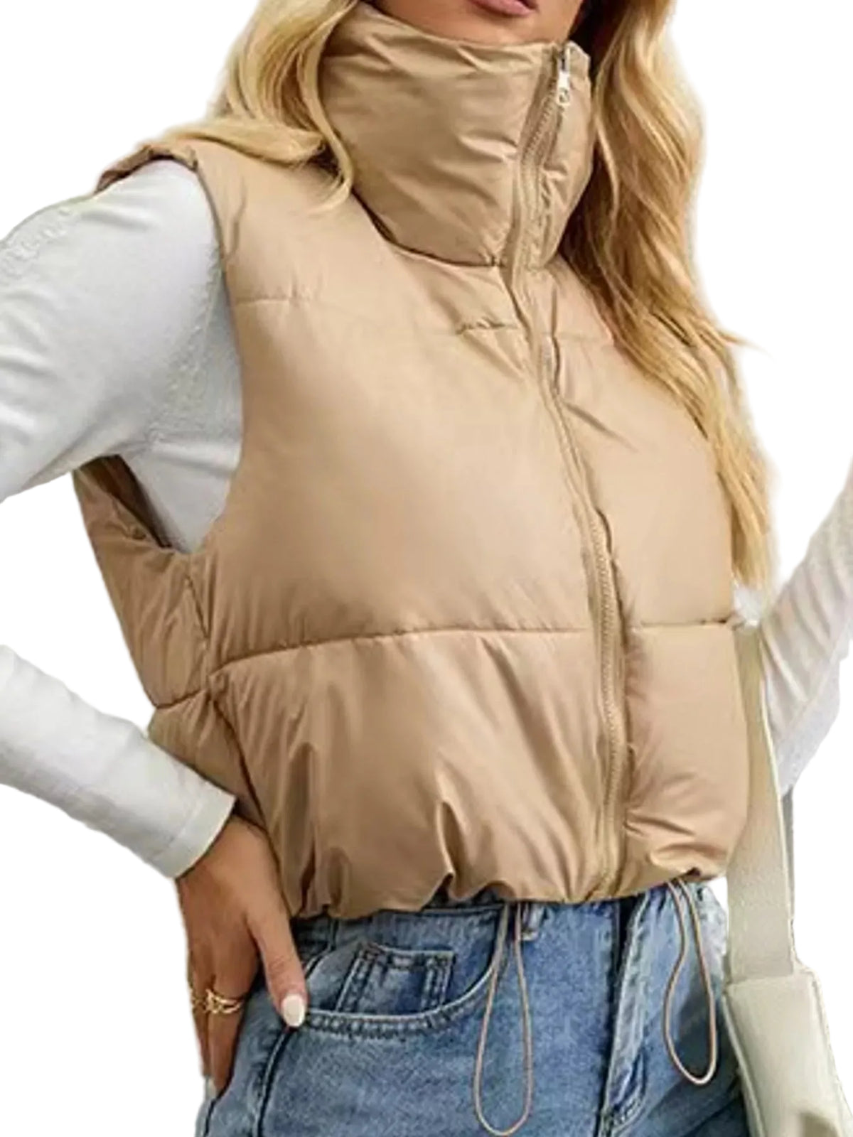 A woman wearing a beige quilted puffer vest over a white long-sleeve top and blue jeans, standing against a textured white wall.