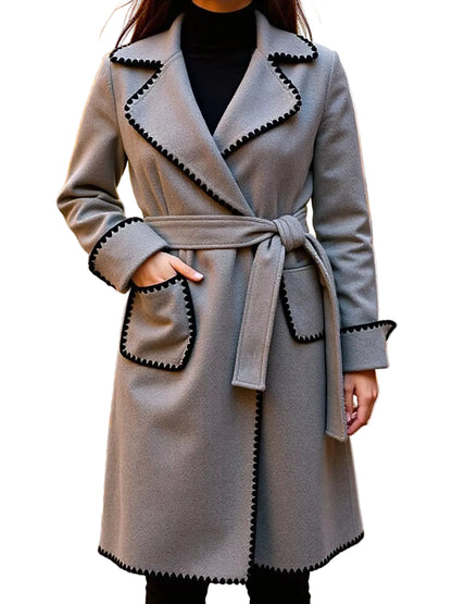 A woman wearing a light gray belted coat with black scalloped trim, photographed outdoors on a street with fallen leaves and blurred buildings in the background.