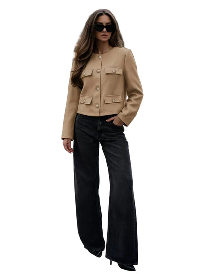 A woman stands on a city crosswalk wearing a cropped tan jacket with gold-tone buttons and flap pockets, paired with wide-leg black jeans and black pointed shoes.