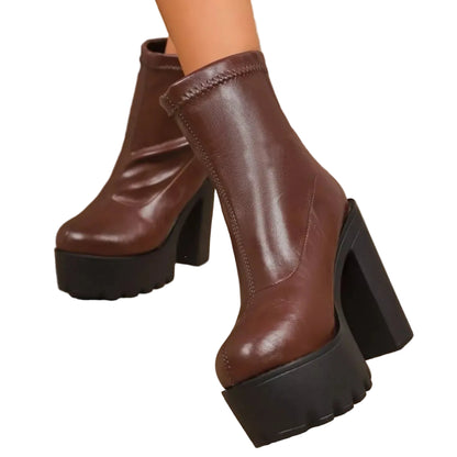 A pair of women's brown faux-leather ankle boots with chunky black platform heels, shown on a model's feet against a warm wood and tile background.