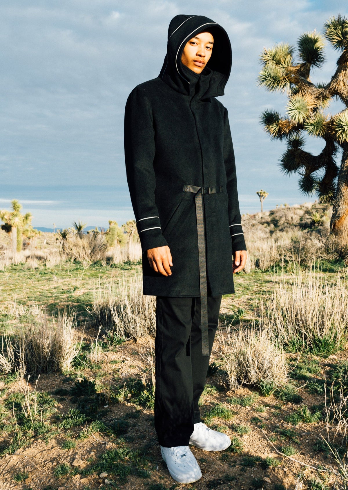 A model wearing a black hooded coat with white stripe details on the cuffs, black pants, and white sneakers, standing in a desert landscape with Joshua trees.