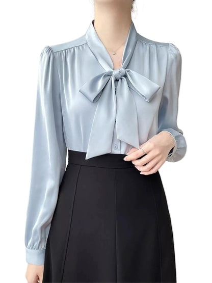A woman wearing a light blue satin blouse with a large bow at the collar and a black skirt, shown from the neck down against a plain light background.
