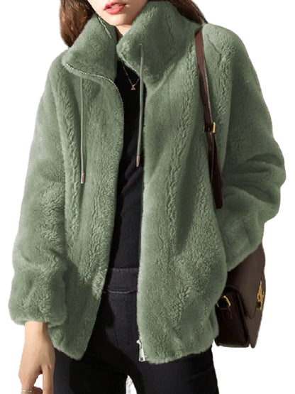 A woman wearing a plush sage green teddy fleece jacket over a black top, carrying a brown shoulder bag against a textured gray wall.