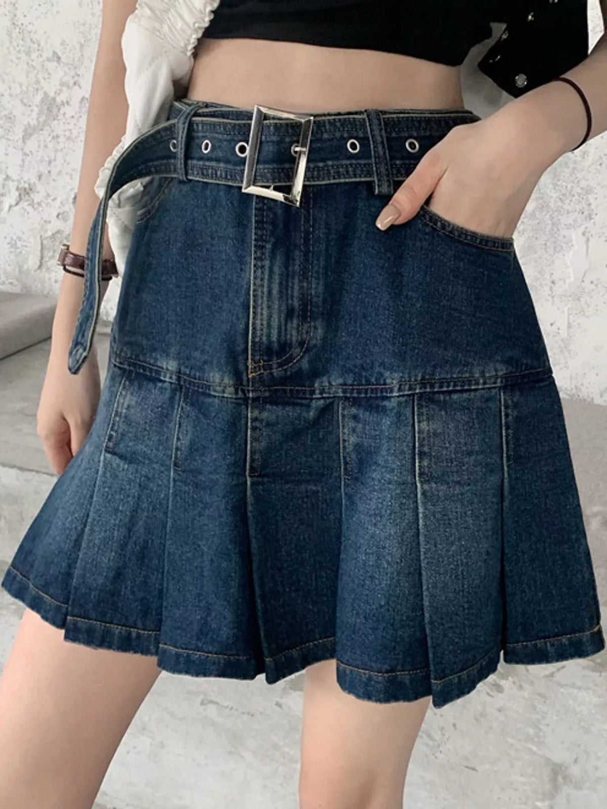A dark blue denim pleated mini skirt with a matching belt featuring a silver buckle, shown on a model.