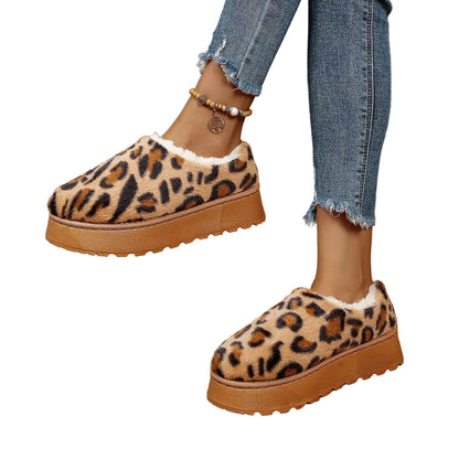 A person wearing leopard-print slip-on platform slippers with white faux-fur lining, paired with frayed-hem blue jeans, standing on a brown tiled floor.