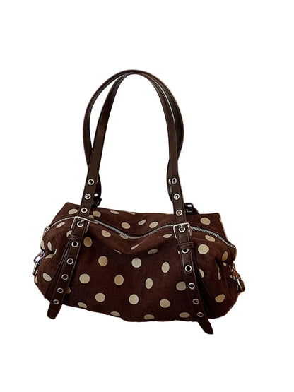 A brown suede handbag with cream polka dots and dark brown leather straps, resting on a wooden floor against a white paneled wall.