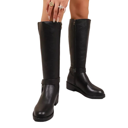 A pair of knee-high black faux-leather boots with side zippers and ankle straps, shown on a model's feet against a beige wall and white marble floor.