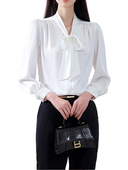 A woman wearing a white long-sleeve blouse with a large front bow and black pants, holding a small black crocodile-embossed handbag with gold hardware.