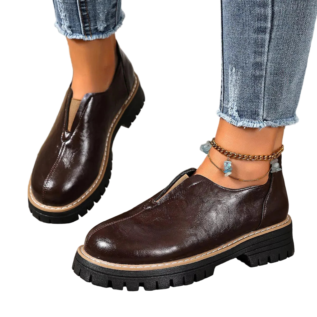 A pair of dark brown slip-on platform shoes with chunky black lug soles, shown on a model wearing blue jeans and ankle bracelets on a rustic wooden deck.