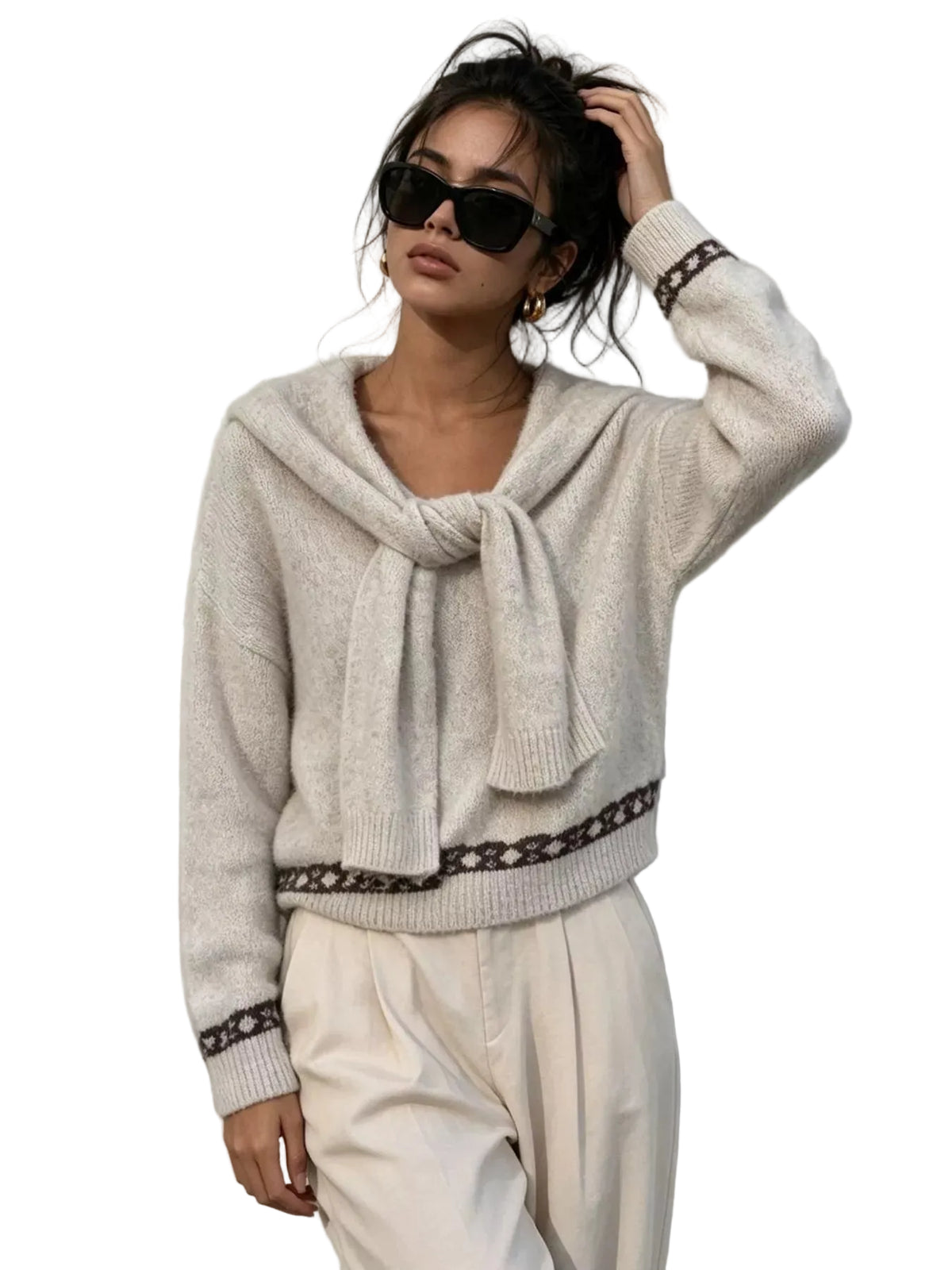A woman wearing a cream knit sweater with a large front bow and dark patterned trim, paired with cream pants, standing against a textured white wall.