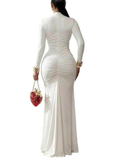 A woman viewed from behind wearing a fitted white long-sleeve mermaid gown with dramatic back ruching and a flared hem, standing in a bright white architectural corridor. She is holding a small red heart-shaped handbag with gold hardware.