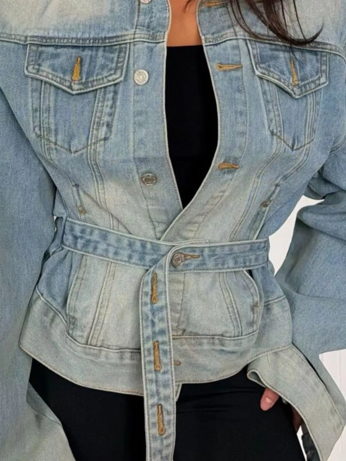 A close-up of a person wearing a light-wash blue denim jacket with a belted waist, layered over a black top and black bottoms.