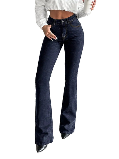 A person wearing dark blue flare jeans with a white cropped knit top and black pointed shoes, standing in a minimalist gray room.