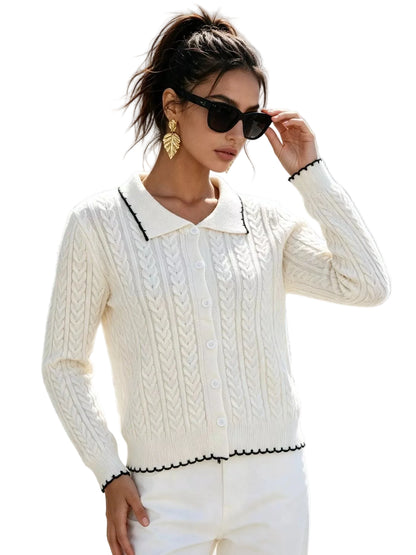 A woman wearing a cream cable-knit button-up cardigan with black trim, paired with white pants and black sunglasses, standing outdoors against a bright, modern building background.