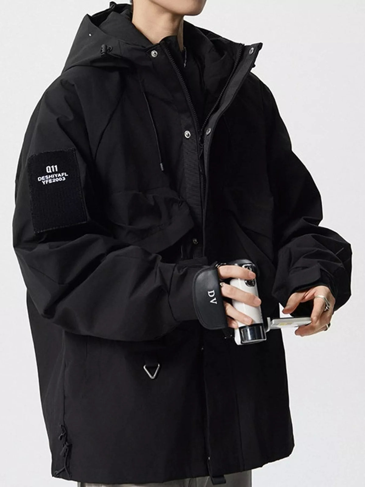 A person wearing a black hooded jacket with a sleeve patch reading 'G111 DESIGN DEPARTMENT' and holding a black and silver tumbler. The jacket has a front zipper, snap buttons, and a drawstring hood, photographed against a light gray studio background.