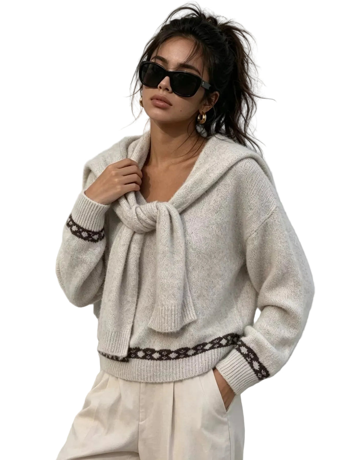 A woman wearing a light beige knit hooded sweater with dark patterned trim, cream pants, and black sunglasses, posing against a textured white wall.
