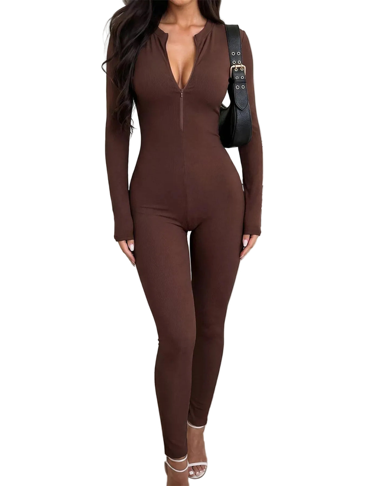 A woman wearing a long-sleeve, form-fitting brown jumpsuit with a deep V-neckline and front zipper, styled with a black shoulder bag and clear high-heeled sandals in a modern indoor setting.