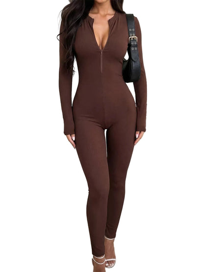 A woman wearing a long-sleeve, form-fitting brown jumpsuit with a deep V-neckline and front zipper, styled with a black shoulder bag and clear high-heeled sandals in a modern indoor setting.