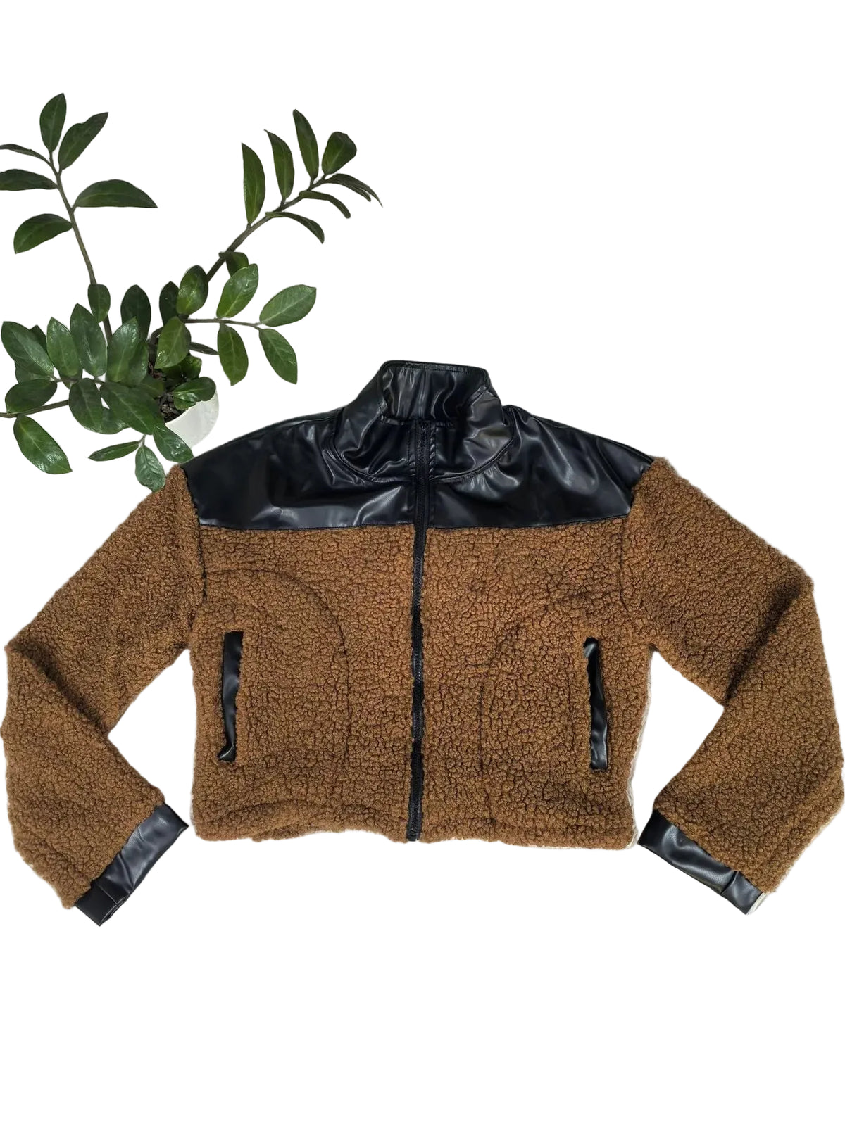 A cropped brown sherpa fleece jacket with black faux-leather panels on the upper chest, collar, and cuffs, laid flat on a white shag rug with a green leafy branch in the corner.
