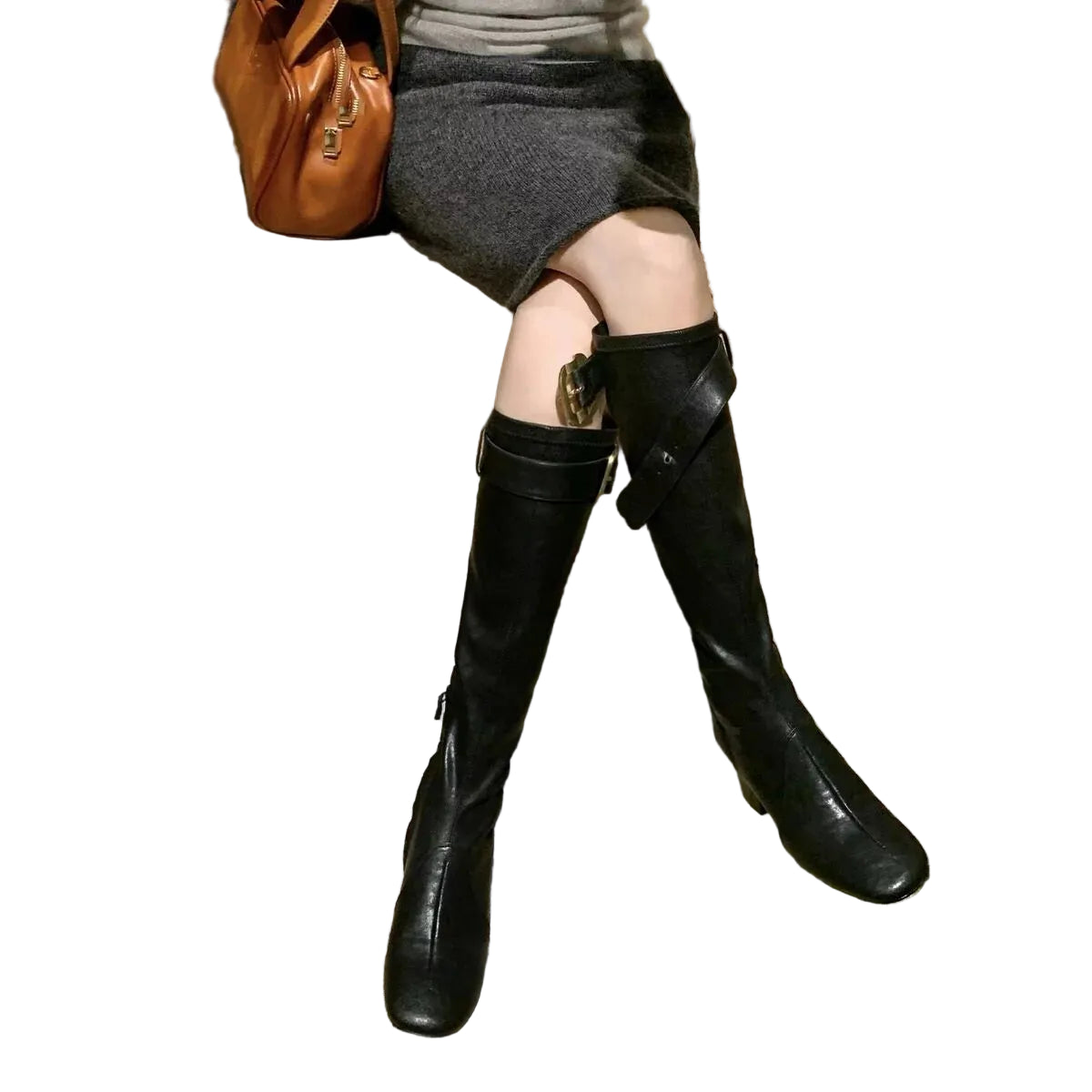 A person seated indoors wearing knee-high black leather boots with a side strap and buckle detail, paired with a gray skirt and carrying a brown leather bag.