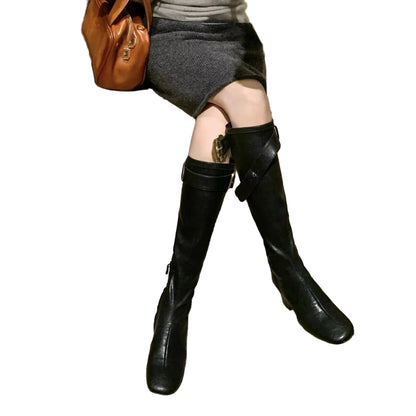 A person seated indoors wearing knee-high black leather boots with a side strap and buckle detail, paired with a gray skirt and carrying a brown leather bag.
