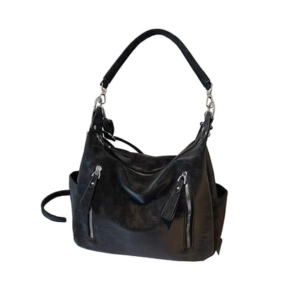 A black leather hobo shoulder bag with silver-tone hardware, displayed on a white book next to a black suitcase against a textured beige wall.