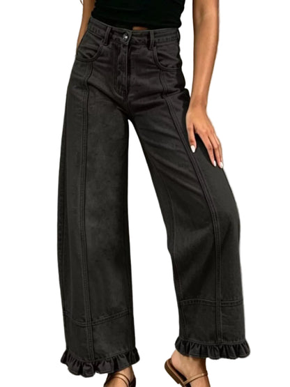 A person wearing high-waisted wide-leg black denim jeans with ruffled hems, paired with a black top and brown sandals, standing against a textured gray wall.