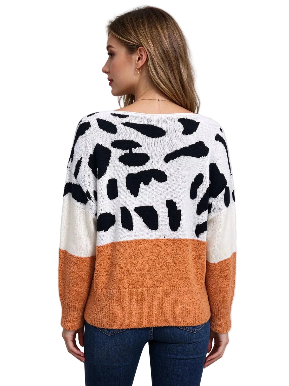 A woman shown from the back wearing a fuzzy knit sweater with a color-blocked design: white upper section with black irregular spots, cream lower left panel, and orange lower right panel and sleeves, paired with blue jeans against a light gray studio background.