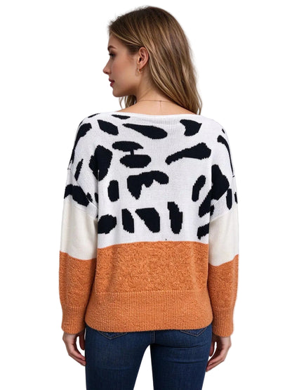 A woman shown from the back wearing a fuzzy knit sweater with a color-blocked design: white upper section with black irregular spots, cream lower left panel, and orange lower right panel and sleeves, paired with blue jeans against a light gray studio background.