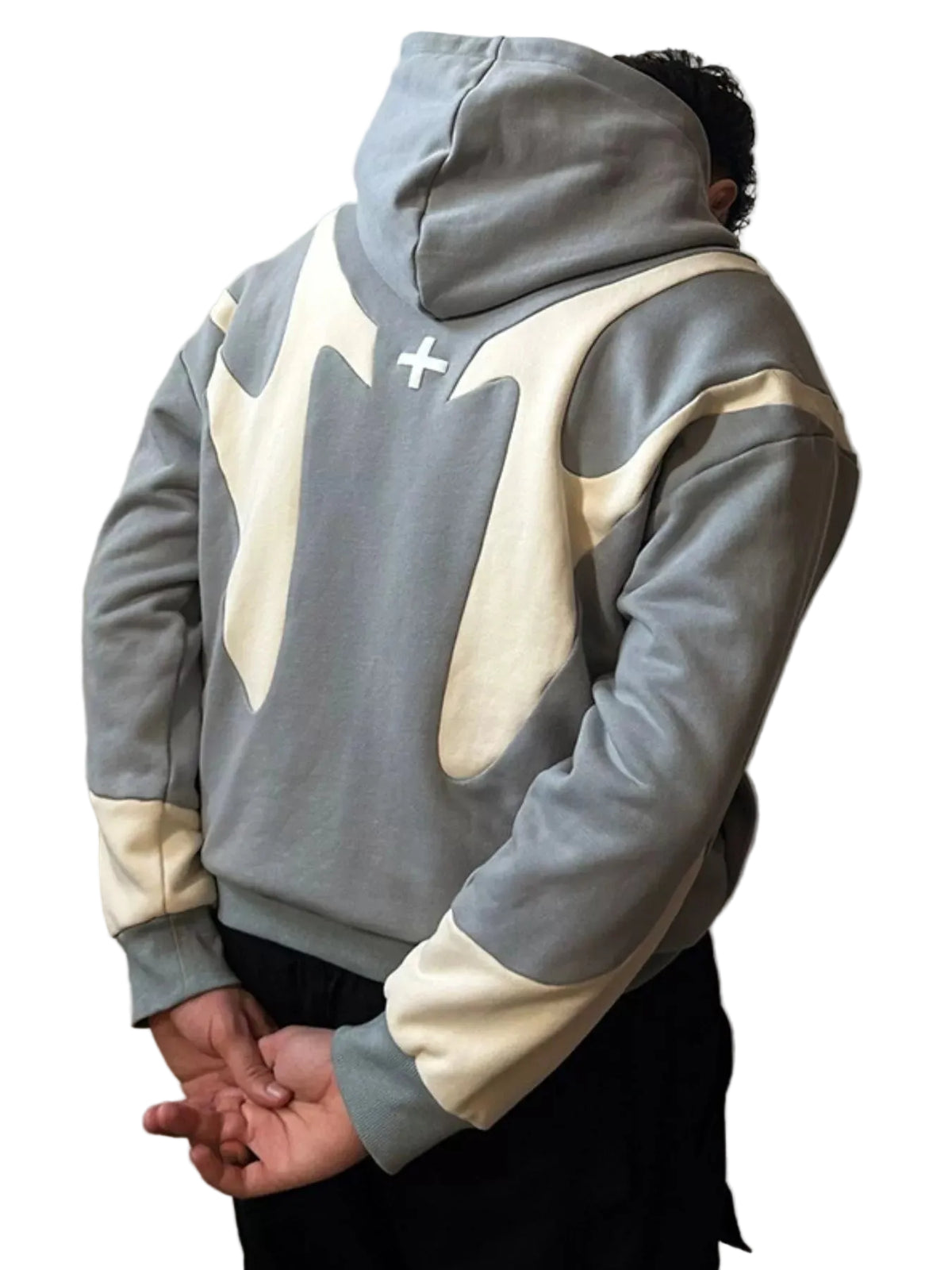 A person shown from the back wearing a gray and cream color-blocked hoodie with a white cross graphic on the upper back, paired with black pants, against a plain light gray wall.