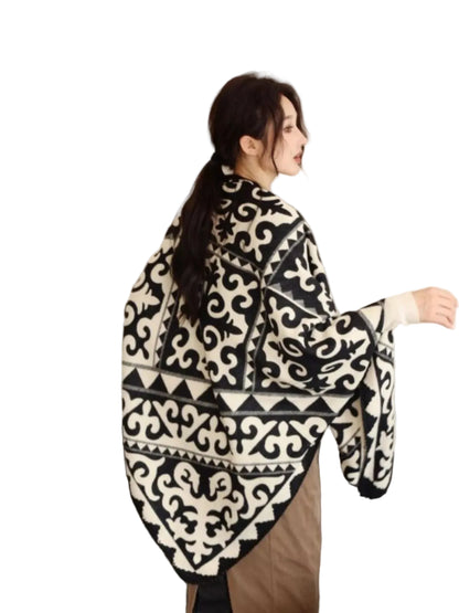 A woman wearing a black and white patterned knit poncho-style cardigan, standing in a living room setting with a dark sofa behind her.