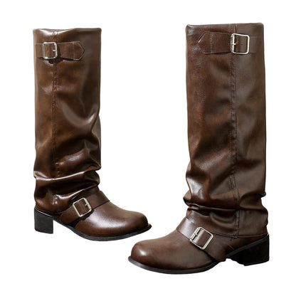 A pair of dark brown leather slouchy knee-high boots with buckle straps, displayed on a dark gray floor against a beige wall and a silver metal block.