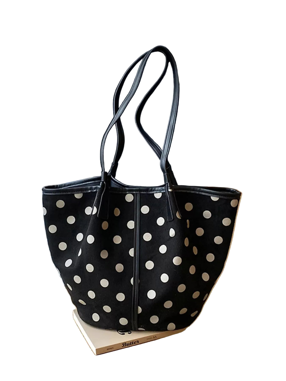 A black canvas tote bag with white polka dots and dark gray shoulder straps, resting on a book on a wooden windowsill with window blinds in the background.