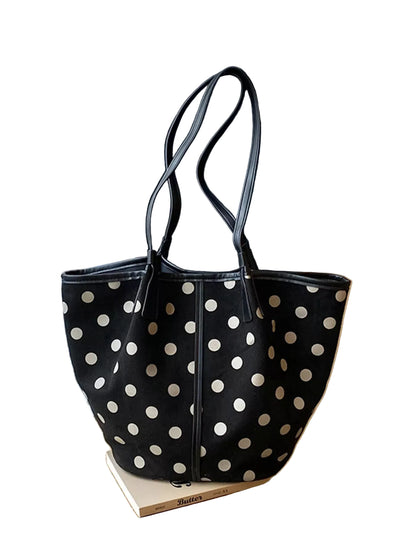 A black canvas tote bag with white polka dots and dark gray shoulder straps, resting on a book on a wooden windowsill with window blinds in the background.