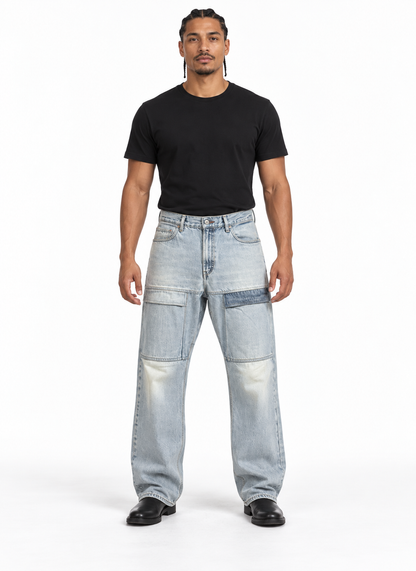 A man modeling a pair of light-wash, wide-leg patchwork jeans with a black t-shirt and black shoes, standing against a plain white studio background.
