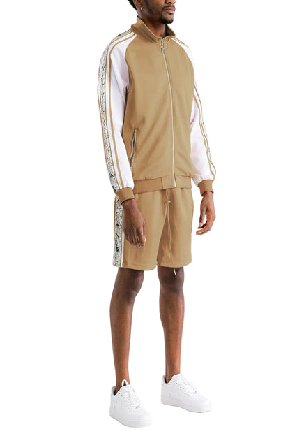 A man modeling a tan and white tracksuit with patterned side stripes, featuring a zip-up jacket and matching shorts, paired with white sneakers against a white studio background.