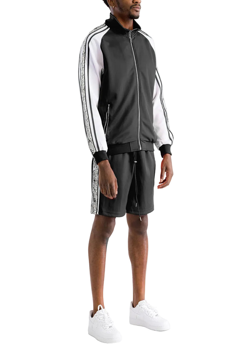 A man modeling a black and white track jacket and matching shorts set with white sneakers, photographed against a plain white studio background.