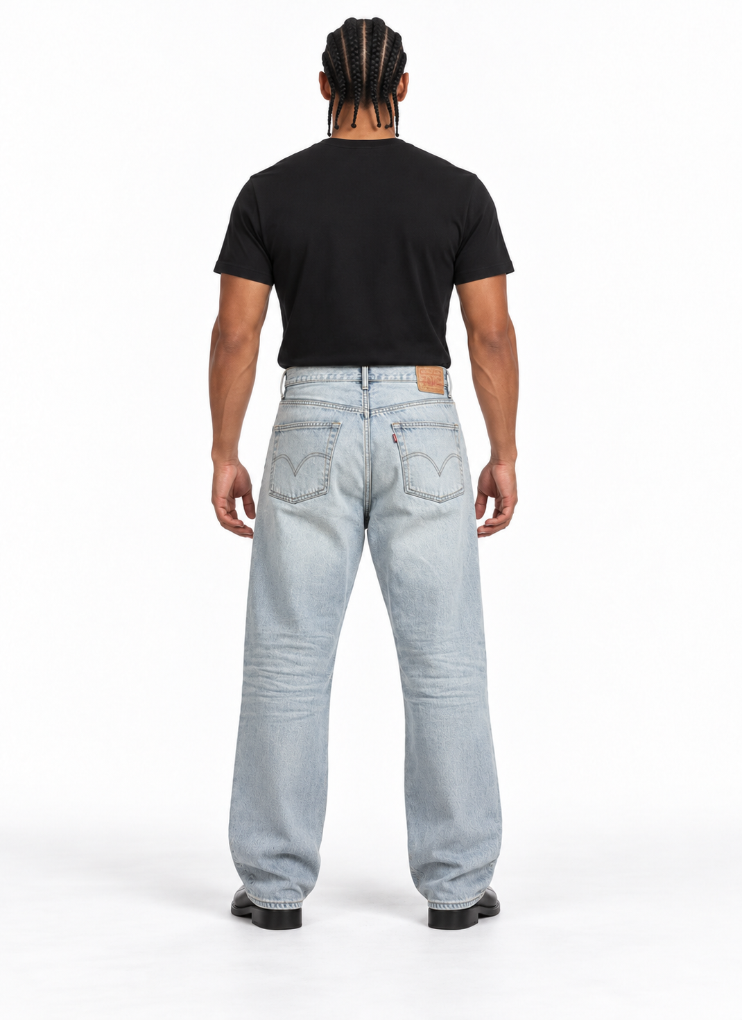 A man viewed from behind wearing a black t-shirt and light-wash blue jeans, standing against a plain white studio background.