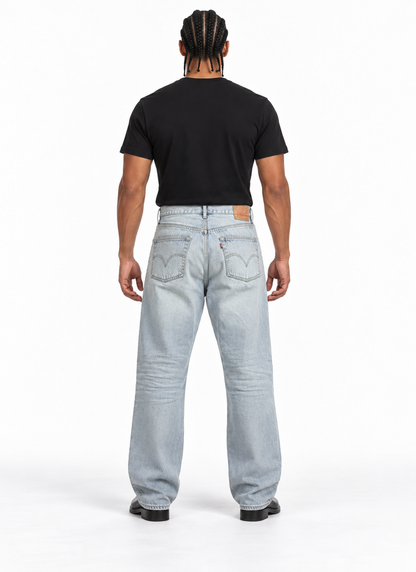 A man viewed from behind wearing a black t-shirt and light-wash blue jeans, standing against a plain white studio background.